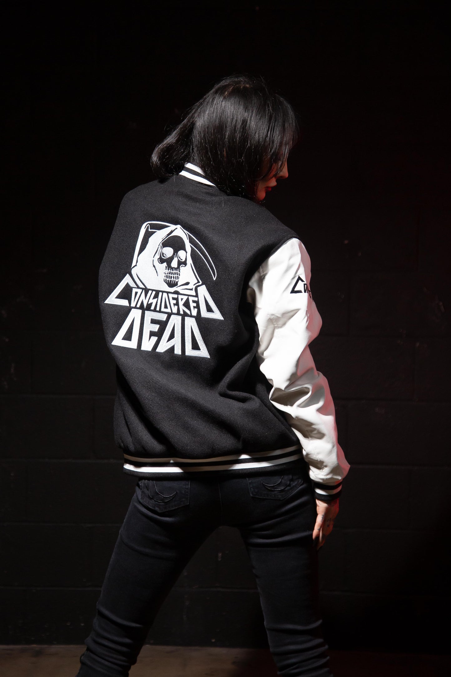 A letterman jacket with a black wool body and white faux leather sleeves. It features the 'Reaper' embroidered on the upper left chest and the words 'Considered Dead' beneath it on the back.