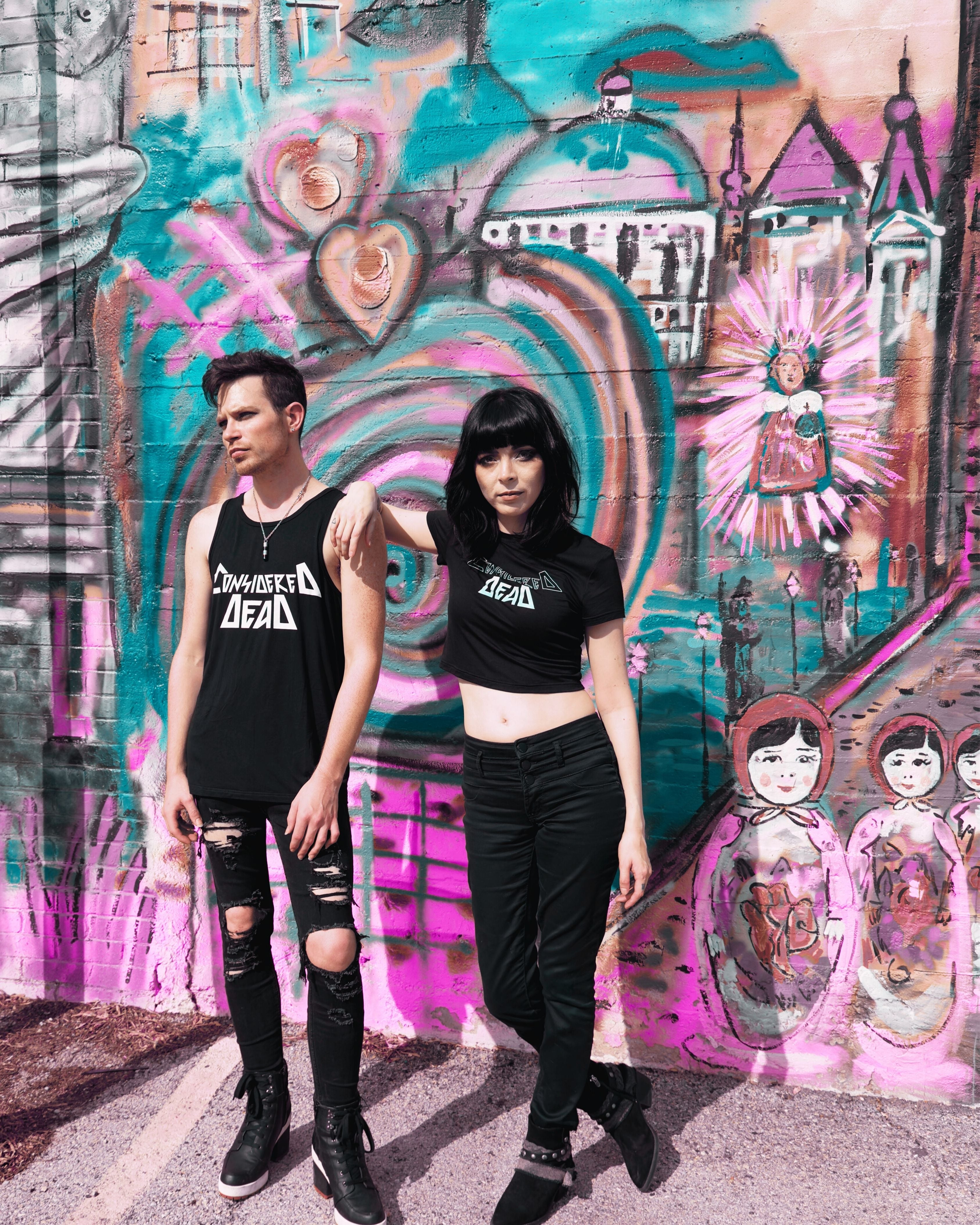 Models wearing Considered Dead sustainable streetwear tank top and crop top in front of a vibrant graffiti wall. Alternative punk and metal-inspired fashion from Dallas, Texas.