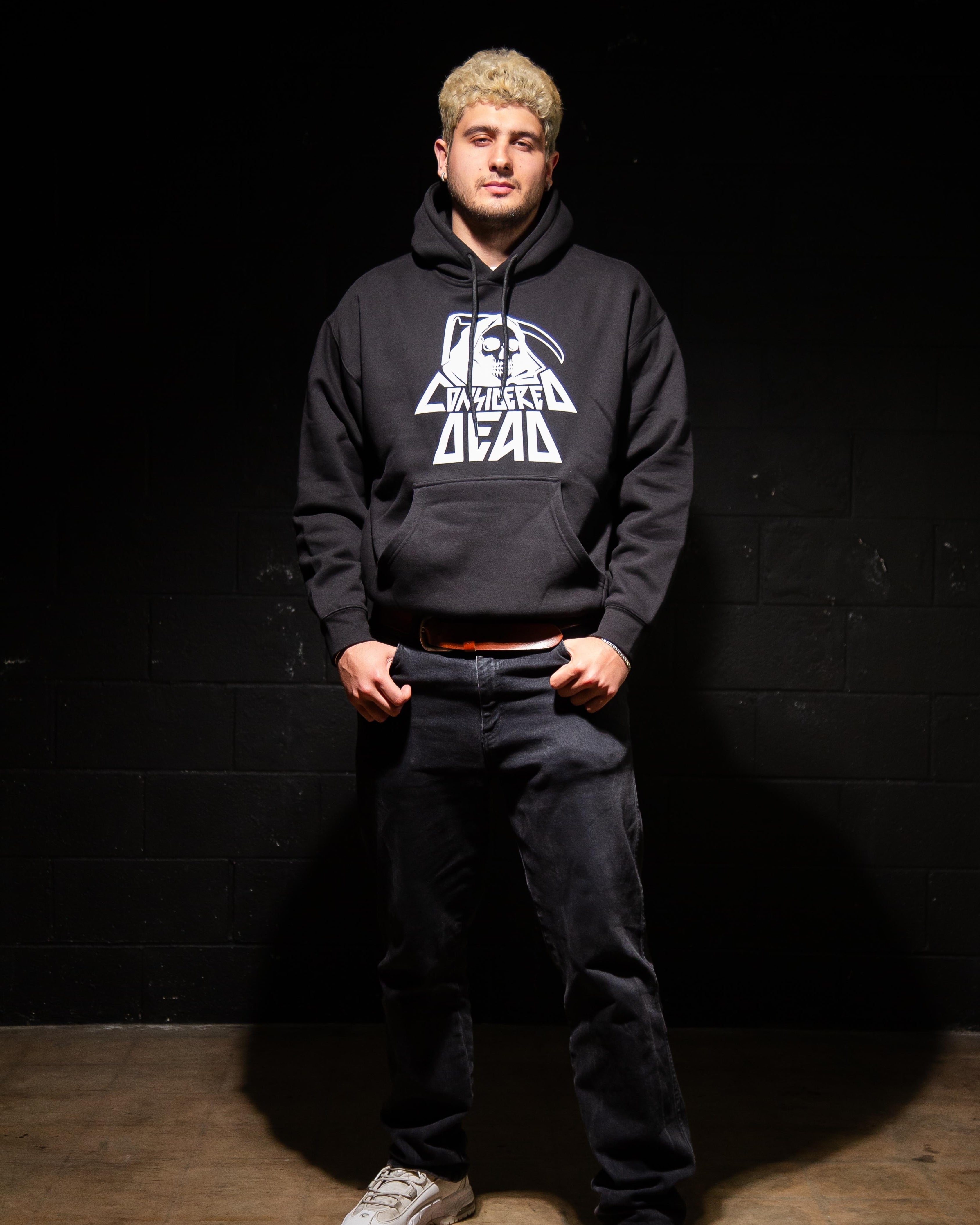 Man wearing a black Considered Dead hoodie with a graphic design against a dark wall.