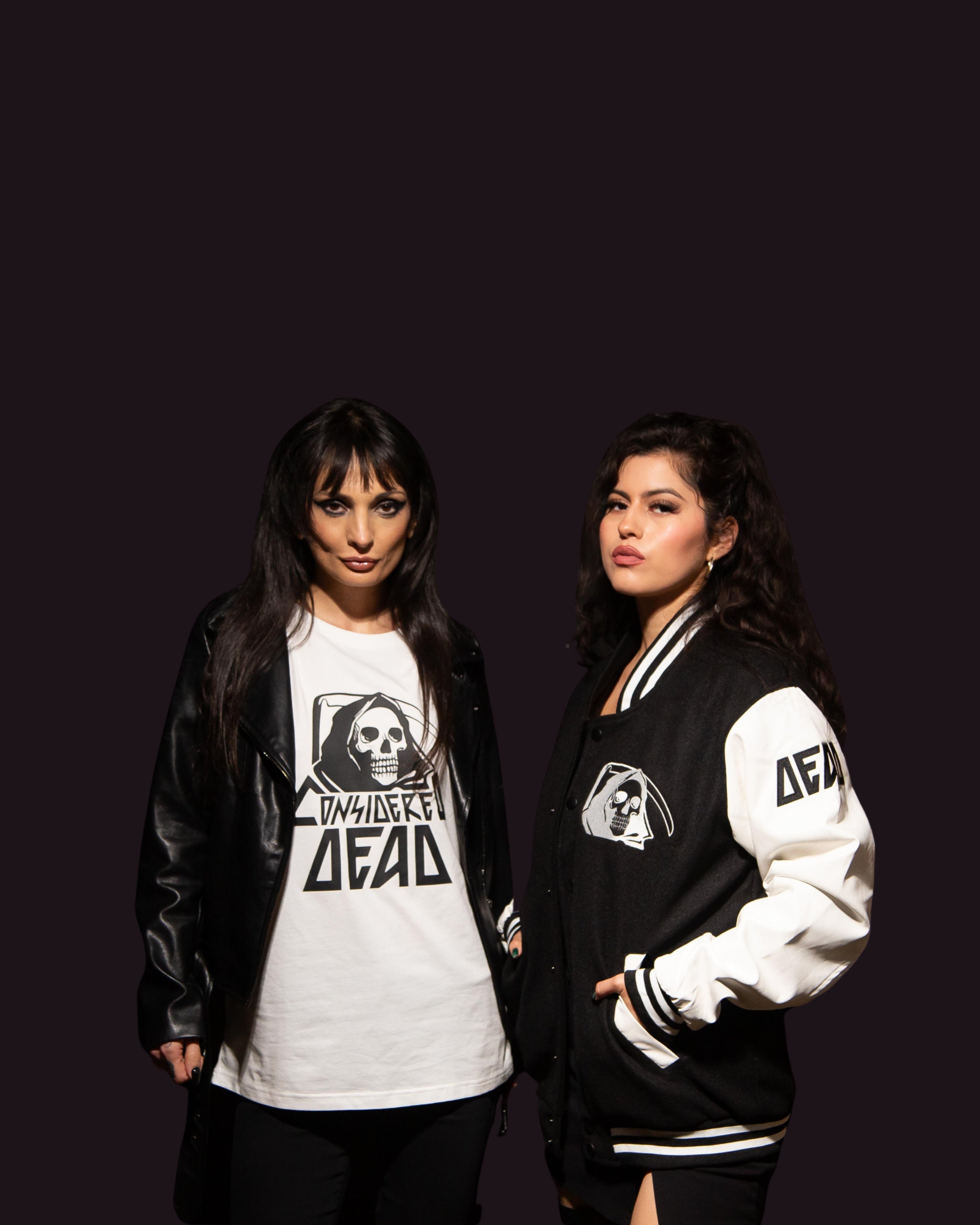 Two women wearing Considered Dead clothing against a dark background. Wearing the white logo tee and Lettermen jacket