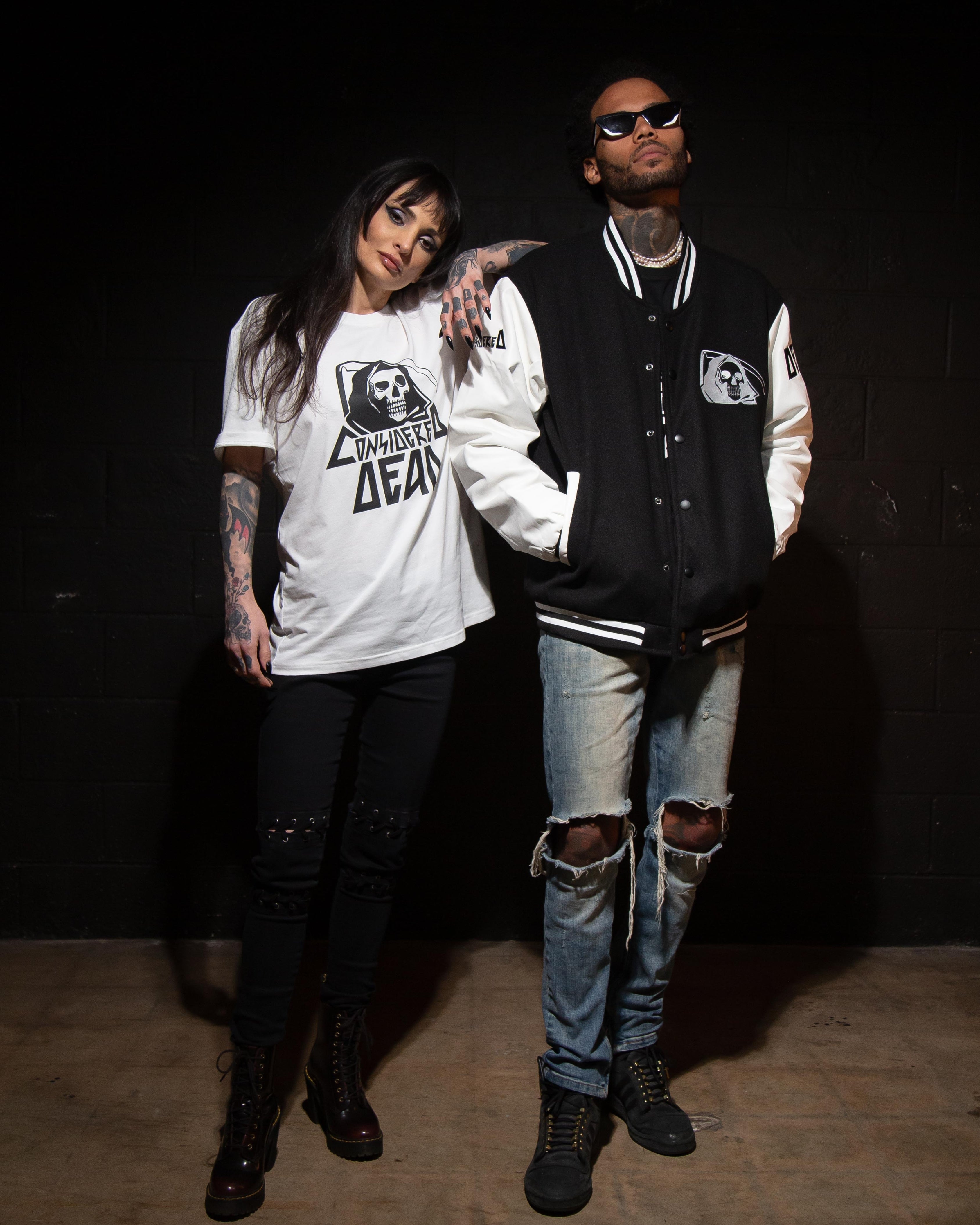 Two people standing against a black background, one wearing a white considered dead t-shirt with a graphic design and the other in a black and white letterman jacket.